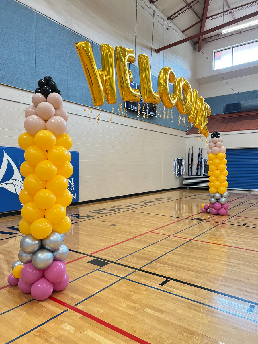 Gold marquee Welcome sign over yellow, pink and black balloon columns in a school gym
